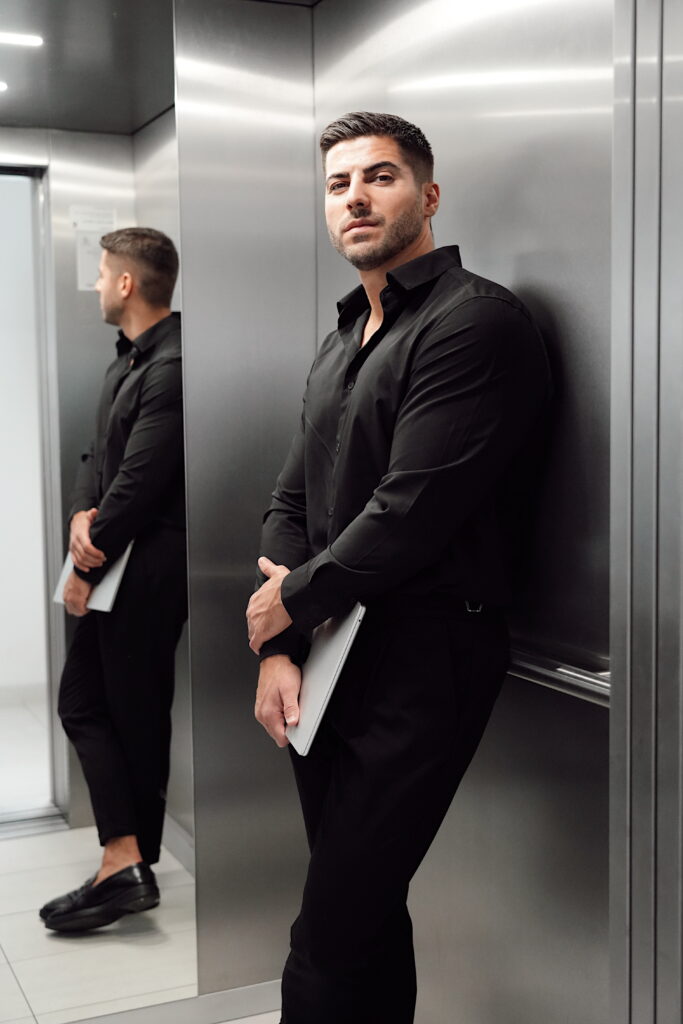 Professional man in black attire holding a laptop in a modern elevator, representing the leadership and expertise behind a Vienna-based marketing agency.