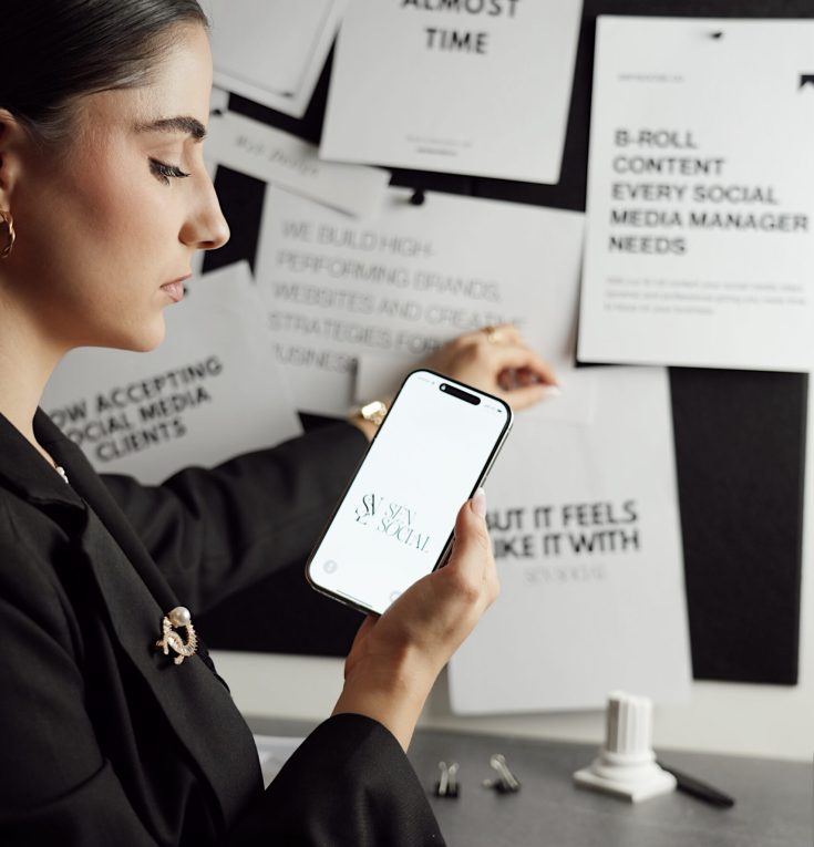 Professional woman holding a smartphone with SÉN Social logo in a creative workspace, surrounded by marketing visuals and brand strategy notes.